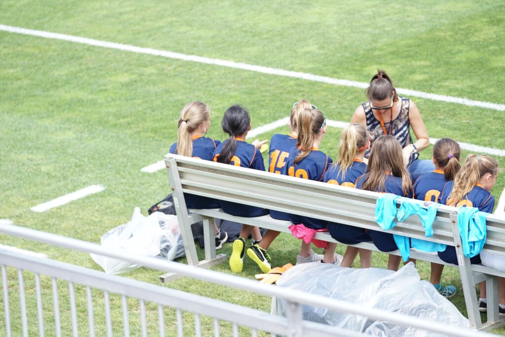 A female coach talking to her team who are sitting on a bench on a soccer field. There are eight young female players in the team who are wearing blue shirts with different numbers on their backs.